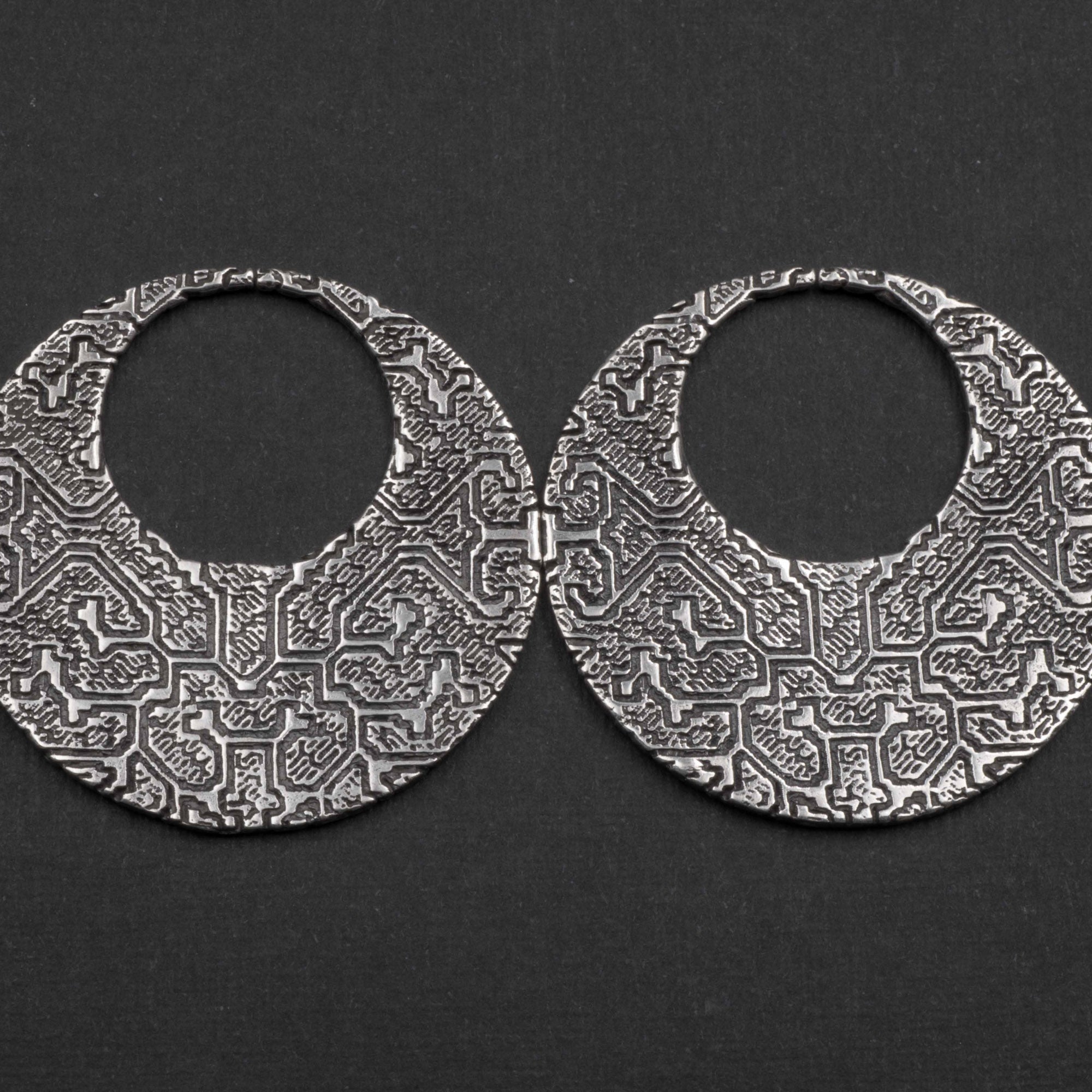 A pair of OLIVIA Tribal Shipibo Seamless Disc Hoop Earrings from Kaya Movement lies on a dark background. These intricately engraved, circular silver earrings feature detailed geometric patterns and textures, lending an antique, handcrafted appearance to the boho statement piece. The 12 gauge earrings are open-hoop in style.